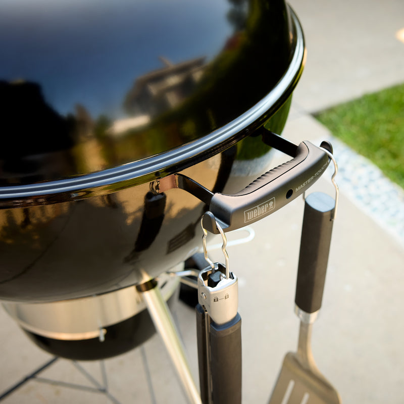 Close-up of a kettle grill’s bowl handle with tongs and a spatula hanging from the two built-in tool hooks.
