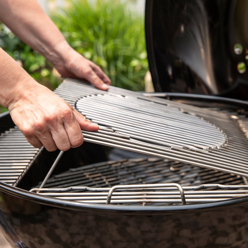 Close-up of hands lifting the cooking grate section with a removable circular center from the Weber 26” Master-Touch Charcoal Grill, revealing the charcoal grate below.