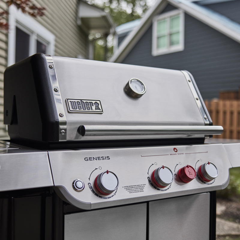 Close-up of a stainless steel Weber Genesis gas grill with a closed lid, built-in thermometer, burner control knobs, and red sear zone knob.
