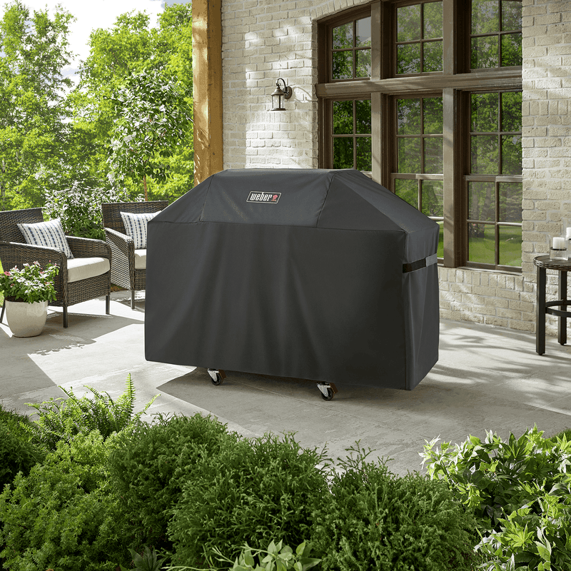 Weber Premium Grill Cover protects a Genesis 3-burner gas grill on a backyard patio surrounded by lush greenery and outdoor seating.