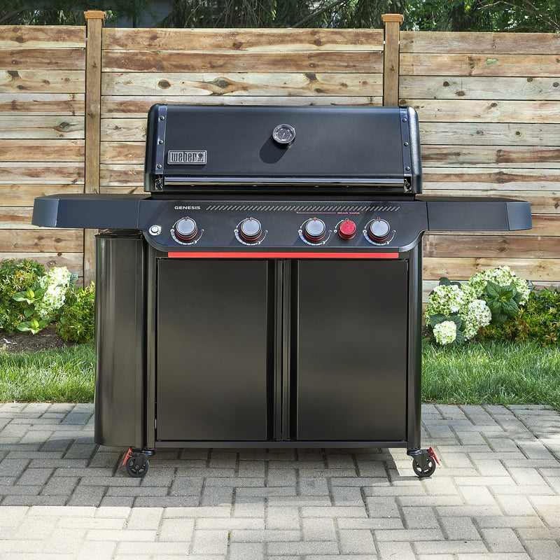 Black Weber Genesis E-430 Stealth Edition gas grill with closed lid and side shelves on a paved patio in front of a wooden fence.