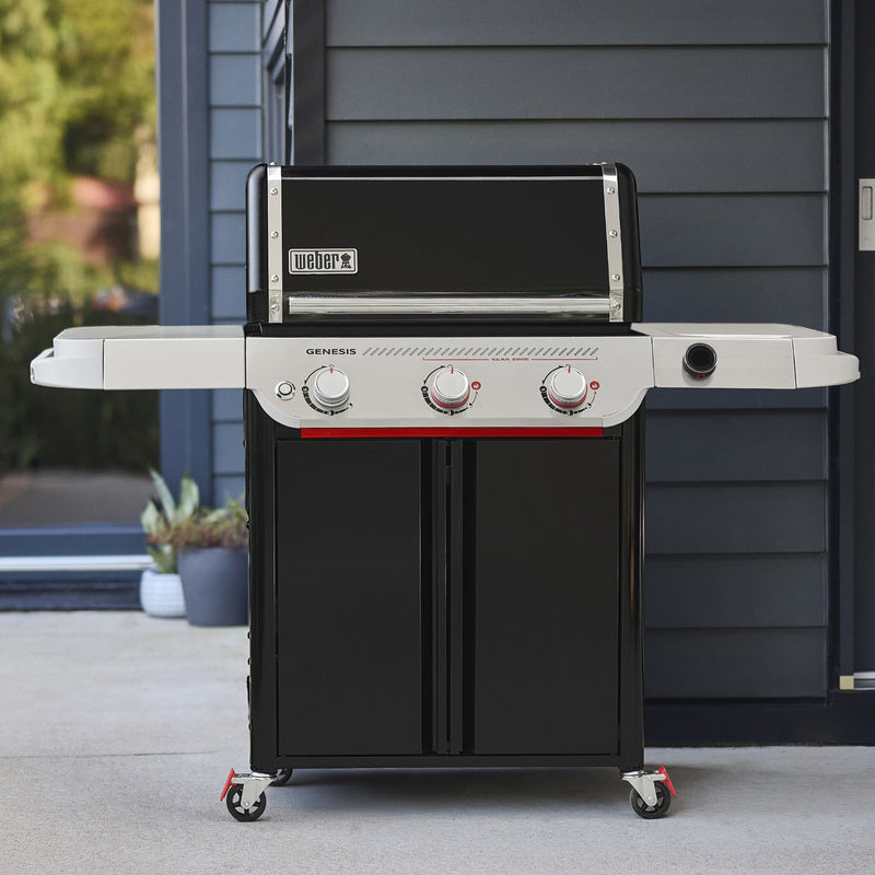 Black Genesis EX-325W Smart Gas Grill on a patio with a closed lid, two side tables, three front control knobs, a digital temperature display, and a two-door cabinet on caster wheels.