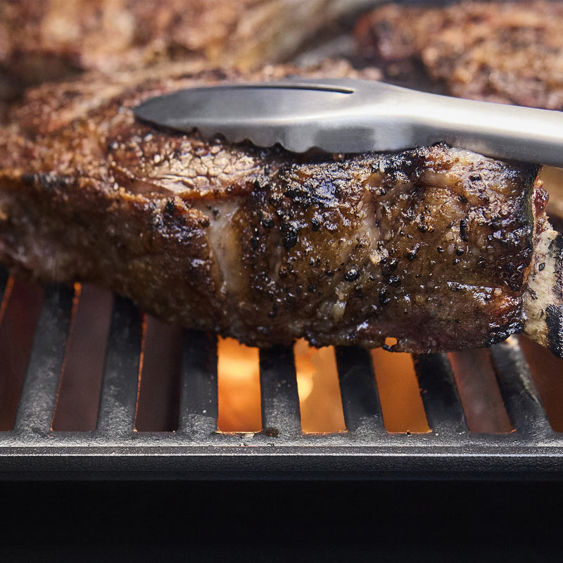 Close-up of metal tongs gripping a seared steak on grill grates with flames below.