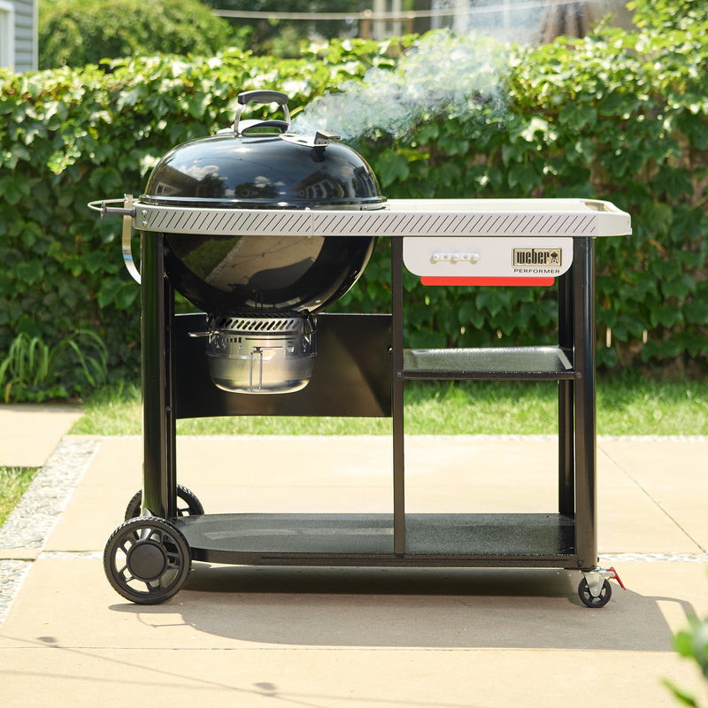 Black Kettle Charcoal Grill on a wheeled cart on a backyard patio, with smoke rising from the closed lid, a large side table, an enclosed ash catcher, and two storage shelves.
