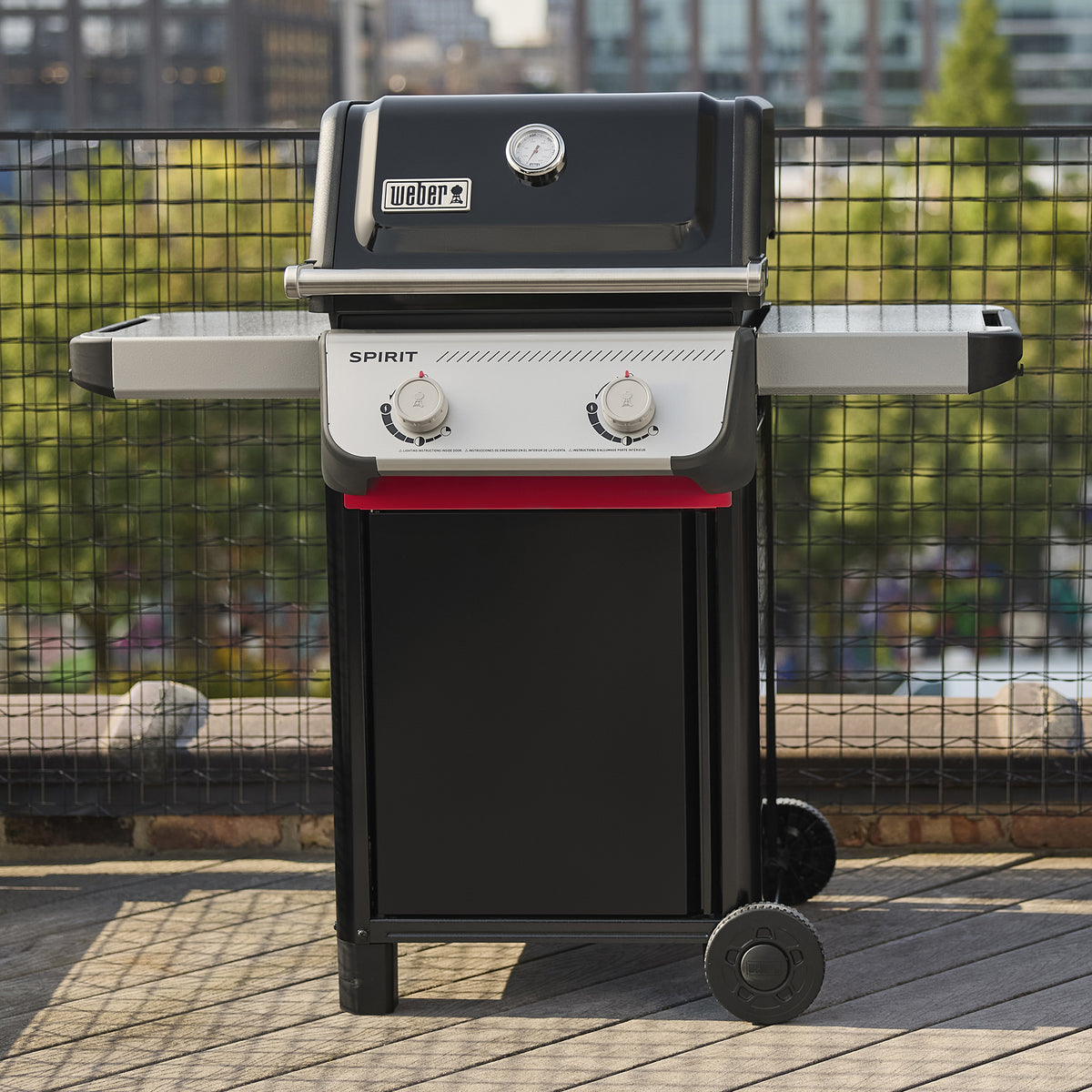 Front view of the black Weber Spirit E-210 Gas Grill on a wooden balcony with the lid closed, two side tables, two control knobs, and city buildings in the background.