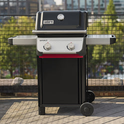 Front view of the black Weber Spirit E-210 Gas Grill on a wooden balcony with the lid closed, two side tables, two control knobs, and city buildings in the background.