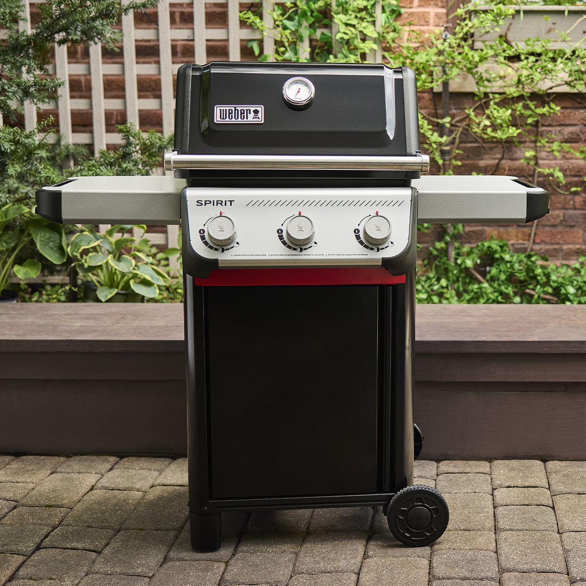 Black Weber Spirit E-310 Gas Grill on a garden patio with the lid closed, a built-in thermometer, two side tables and three control knobs.