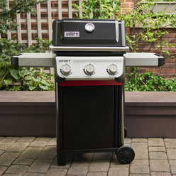Black Weber Spirit E-310 Gas Grill on a garden patio with the lid closed, a built-in thermometer, two side tables and three control knobs.