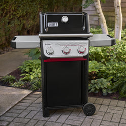 Black Weber Spirit E-325 Gas Grill on a garden patio with the lid closed, two side tables, and three control knobs, including two red-accented Boost Burner knobs.