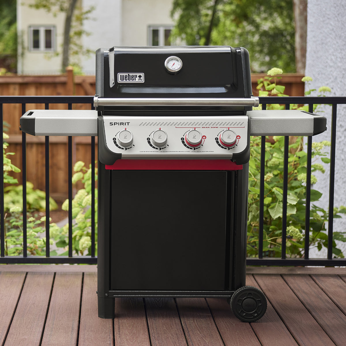 On a wooden deck, the Weber Spirit E-425 Gas Grill has a closed black lid with a built-in thermometer, two side tables, and four burner controls, including two red Boost Burner knobs for the Sear Zone.