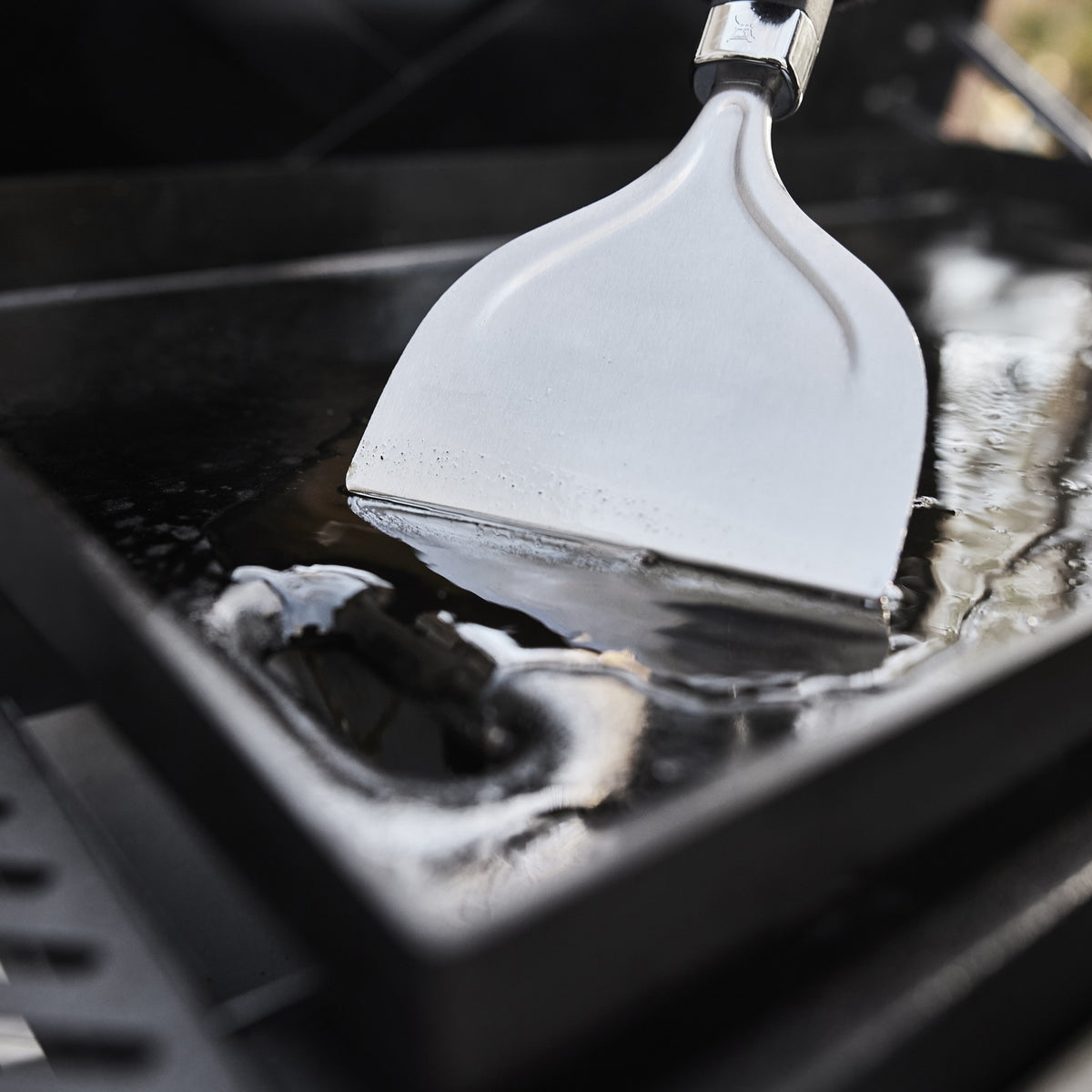 Close-up of a metal scraper pushing liquid across the griddle cooktop toward the front grease opening.