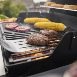 Close-up of a spatula lifting a burger on the Weber Spirit E-210 Gas Grill, with corn and red onion slices cooking alongside, buns on the warming rack, and flames beneath the grates.