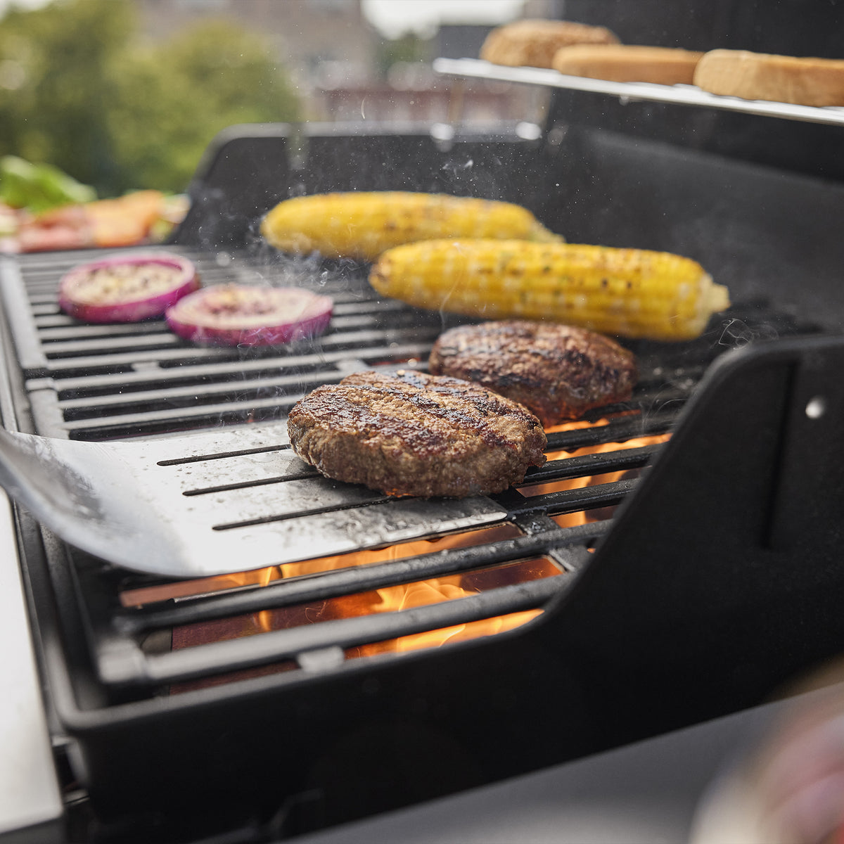 A spatula lifts a burger patty on the Weber Spirit E-310 Gas Grill beside corn and red onion slices, with buns on the warming rack.