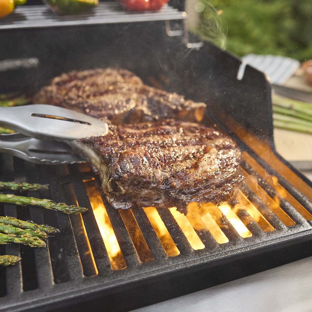 Close-up of tongs gripping a seared steak on the Weber Spirit E-325 Gas Grill, with asparagus alongside and flames beneath the grates.