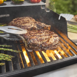 Close-up of tongs gripping a seared steak on the Weber Spirit E-325 Gas Grill, with asparagus alongside and flames beneath the grates.