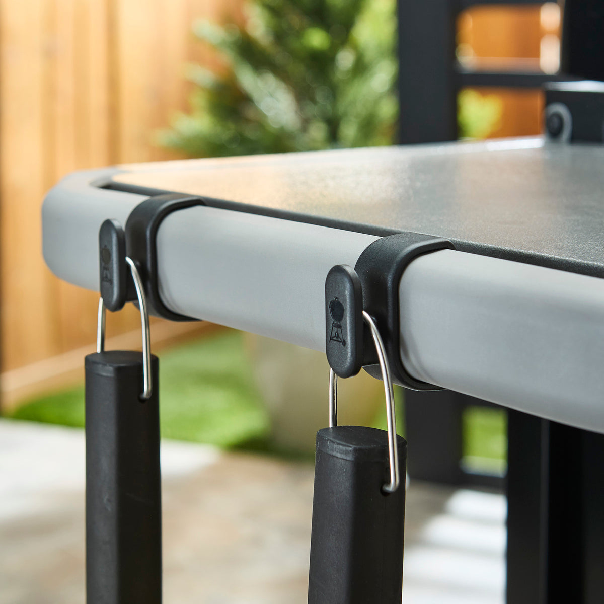 Close-up of two cooking tools hanging from snap-on hooks attached to the griddle’s side table rail.