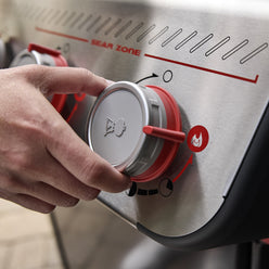 Close-up of a hand adjusting a red-accented Boost Burner knob on the Weber Spirit E-325 Gas Grill beneath the Sear Zone label.