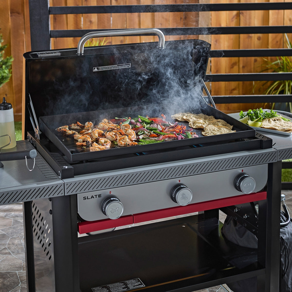 Shrimp, sliced peppers, onions, and tortillas cooking on a griddle with the hard cover open, with a plate of tortillas, lime wedges, and cilantro on the side table.