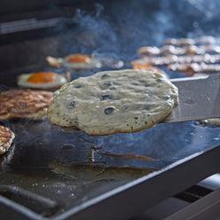 Close-up of a spatula lifting a blueberry pancake from the griddle, with more pancakes, fried eggs, and bacon cooking in the background.
