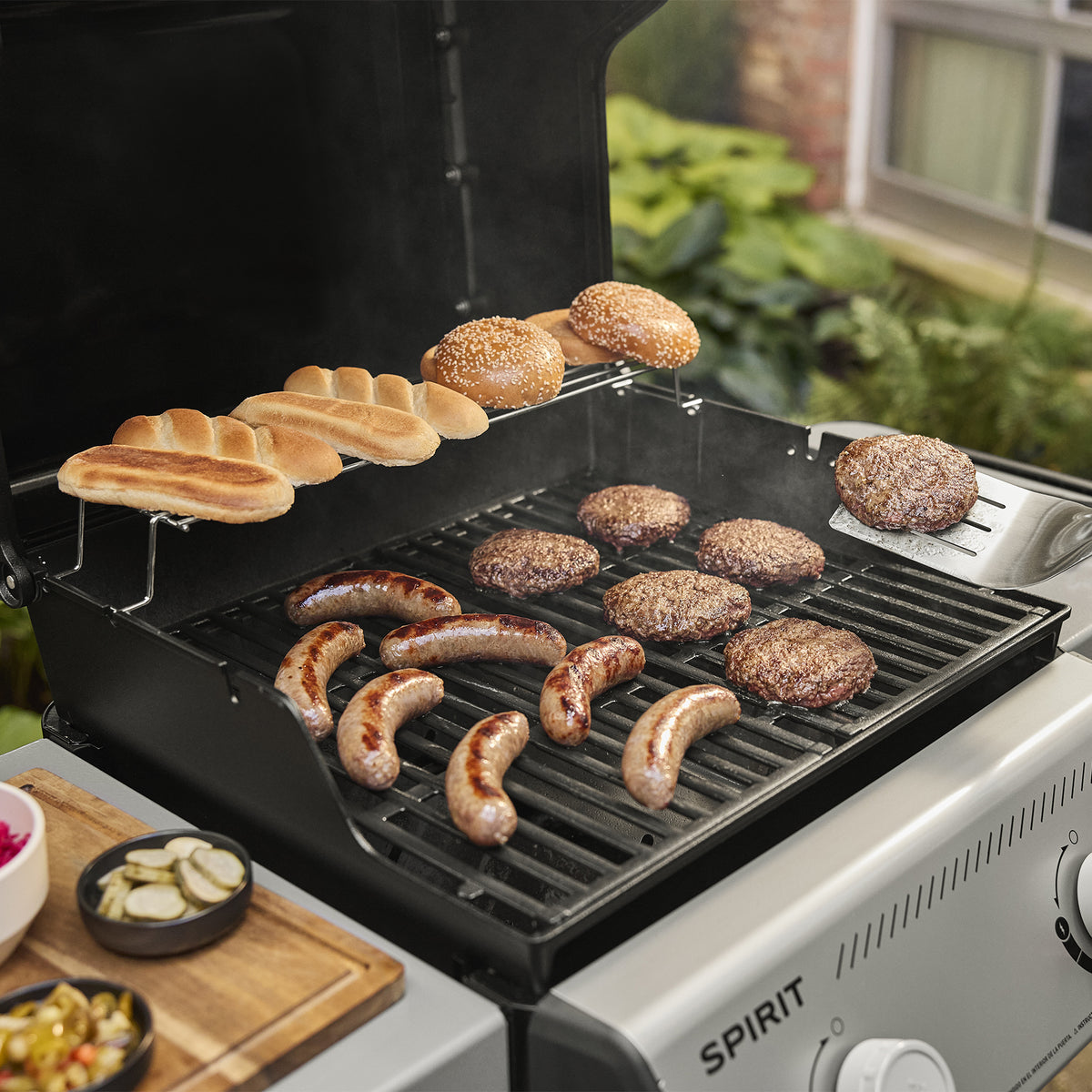 Burgers and sausages cooking on the Weber Spirit E-310 Gas Grill, with buns on the warming rack and a spatula lifting a burger patty.
