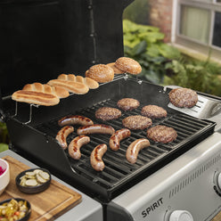 Burgers and sausages cooking on the Weber Spirit E-310 Gas Grill, with buns on the warming rack and a spatula lifting a burger patty.