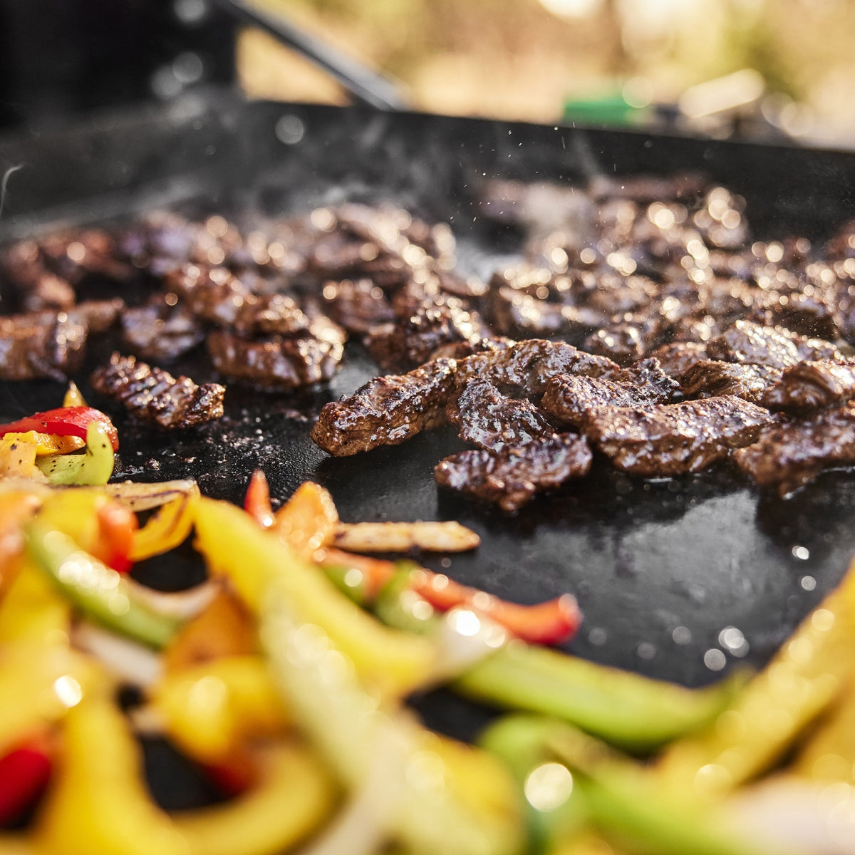 Close-up of seared steak strips, sliced bell peppers, and onions cooking on the griddle.