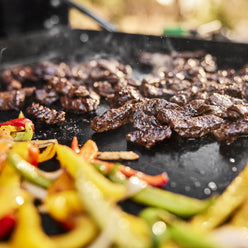 Close-up of seared steak strips, sliced bell peppers, and onions cooking on the griddle.