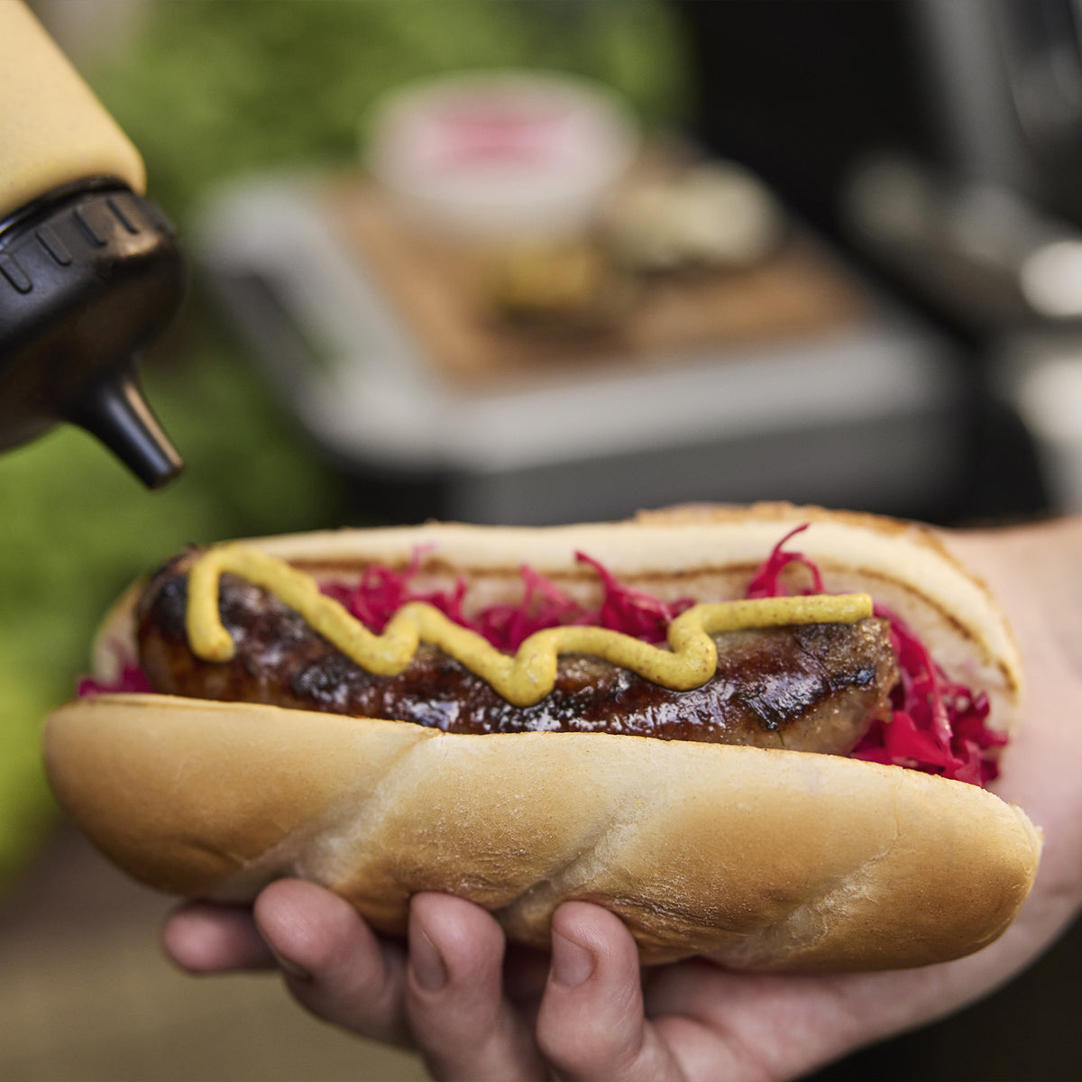 Close-up of a hand holding a grilled sausage in a bun topped with shredded red cabbage and mustard, with the Weber Spirit E-310 Gas Grill in the background.