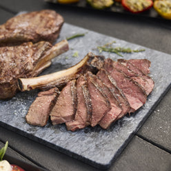 Sliced steak with a browned crust and pink center rests beside whole bone-in steaks on a gray serving board, sprinkled with coarse salt.