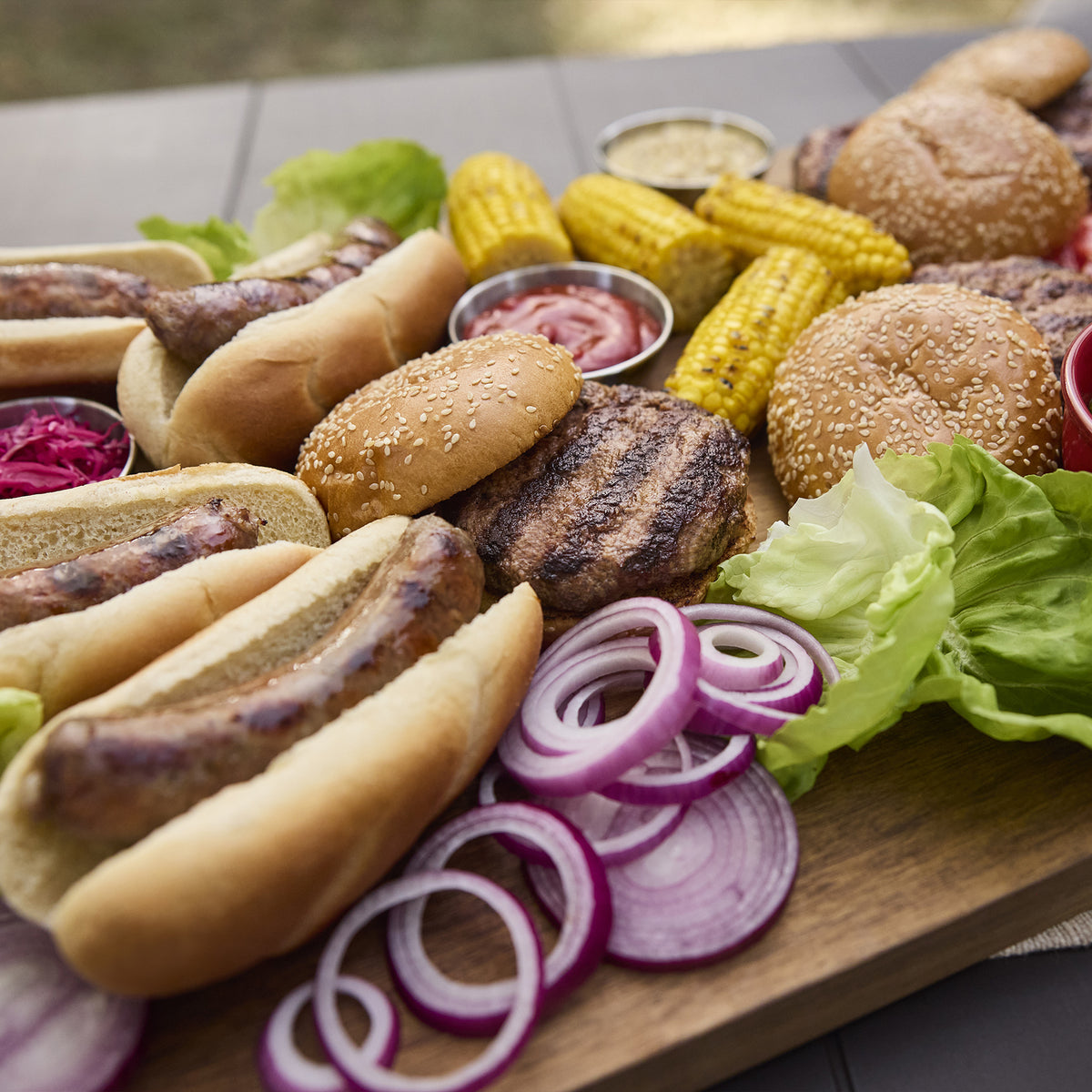 Grilled burgers and sausages in buns on a wooden serving board with corn, lettuce, red onion slices, shredded red cabbage, and condiments.