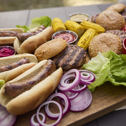 Grilled burgers and sausages in buns on a wooden serving board with corn, lettuce, red onion slices, shredded red cabbage, and condiments.