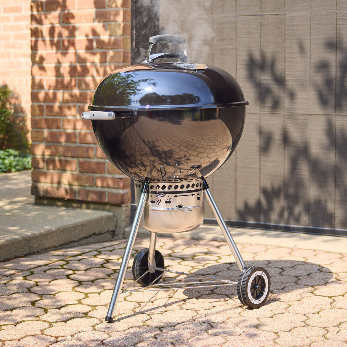 Black Weber 22” Original Kettle Premium charcoal grill cooking with the lid closed as smoke rises on a sunlit stone patio.