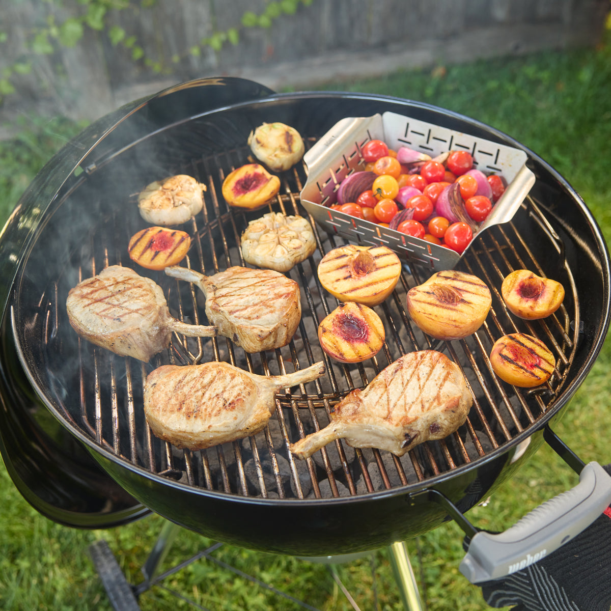 Weber 22” Original Kettle Premium charcoal grill cooks pork chops, peaches, garlic, and a basket of vegetables together.