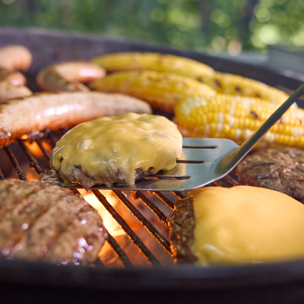 Weber 22” Original Kettle Premium charcoal grill cooks cheeseburgers, sausages, and corn together over hot coals for a backyard cookout.