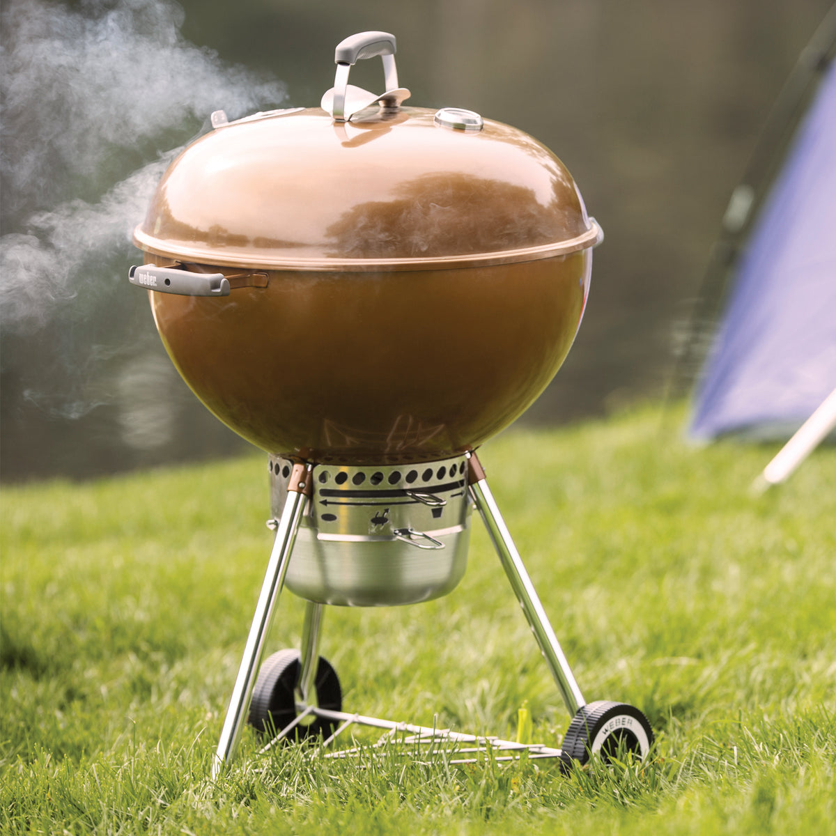 Bronze Weber 22” Original Kettle Premium charcoal grill cooks with the lid closed as smoke rises on a grassy lawn beside a tent.