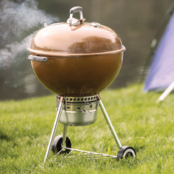 Bronze Weber 22” Original Kettle Premium charcoal grill cooks with the lid closed as smoke rises on a grassy lawn beside a tent.