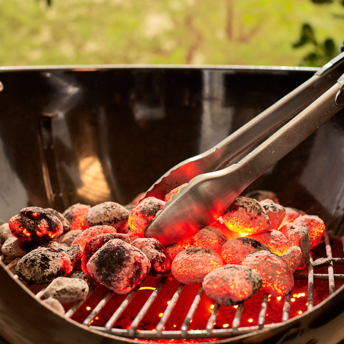 Glowing charcoal is arranged with tongs on the fuel grate of the black Weber 22” Original Kettle Premium charcoal grill to prepare for cooking.
