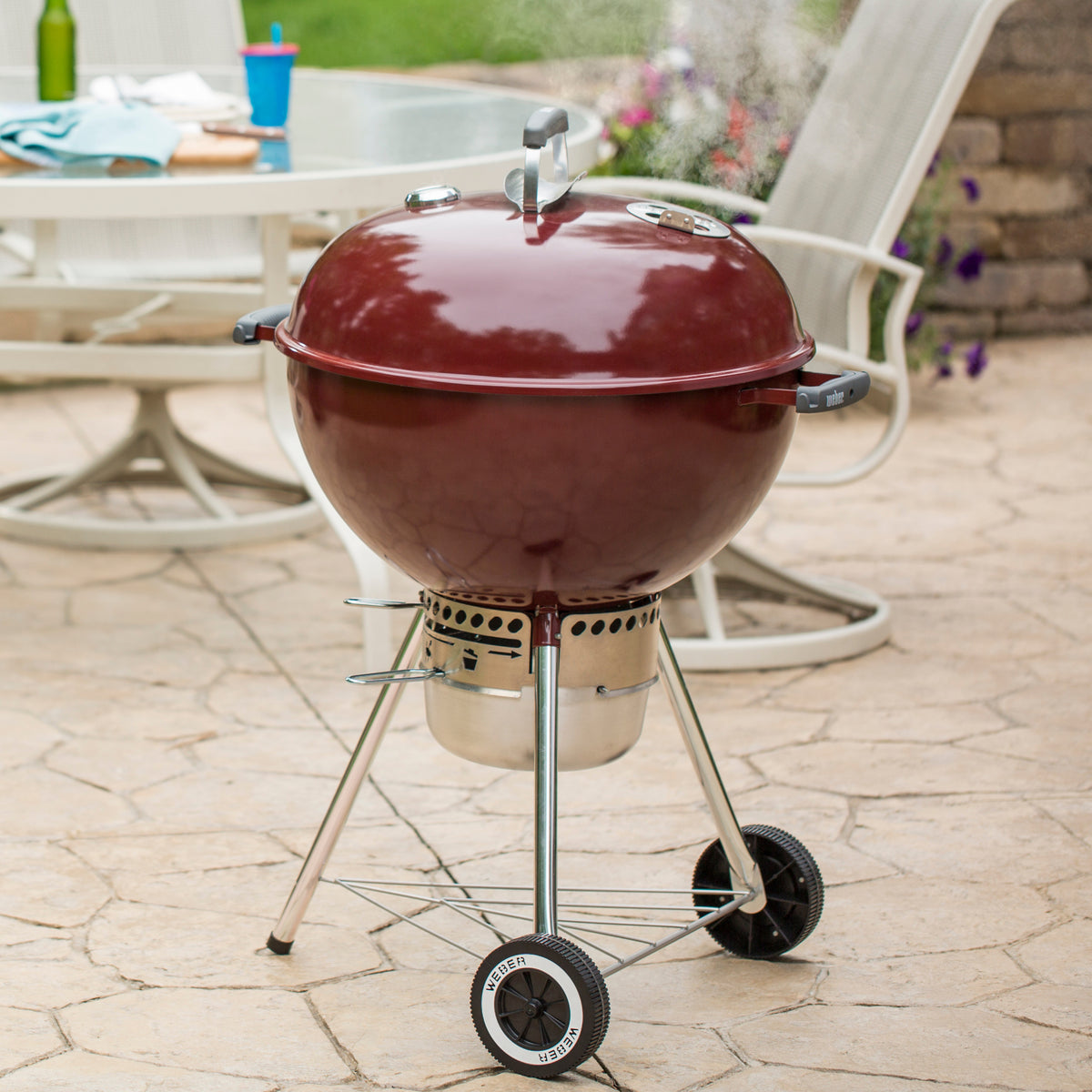 Red Weber 22” Original Kettle Premium charcoal grill cooks with the lid closed on a stone patio beside an outdoor dining table.
