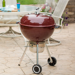 Red Weber 22” Original Kettle Premium charcoal grill cooks with the lid closed on a stone patio beside an outdoor dining table.