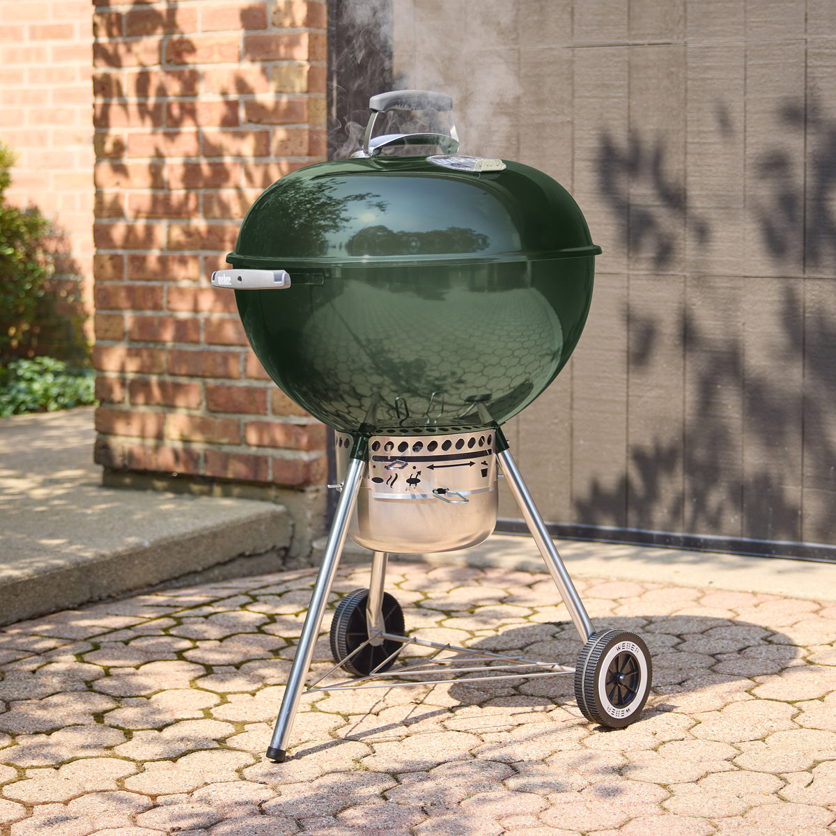 Green Weber 22” Original Kettle Premium charcoal grill cooking with the lid closed as smoke rises on a sunlit stone patio.