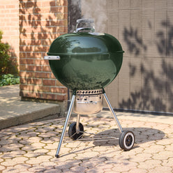 Green Weber 22” Original Kettle Premium charcoal grill cooking with the lid closed as smoke rises on a sunlit stone patio.