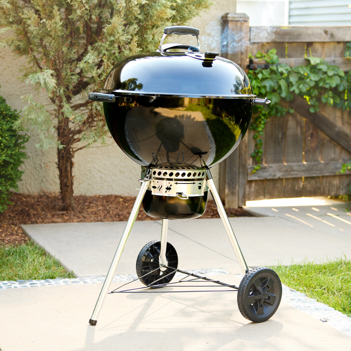 Kettle charcoal grill on a backyard patio with smoke rising from the lid vent, an enclosed ash catcher, two wheels, and a lower wire shelf.