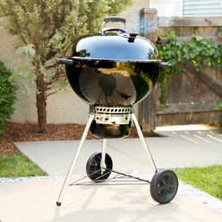 Kettle charcoal grill on a backyard patio with smoke rising from the lid vent, an enclosed ash catcher, two wheels, and a lower wire shelf.