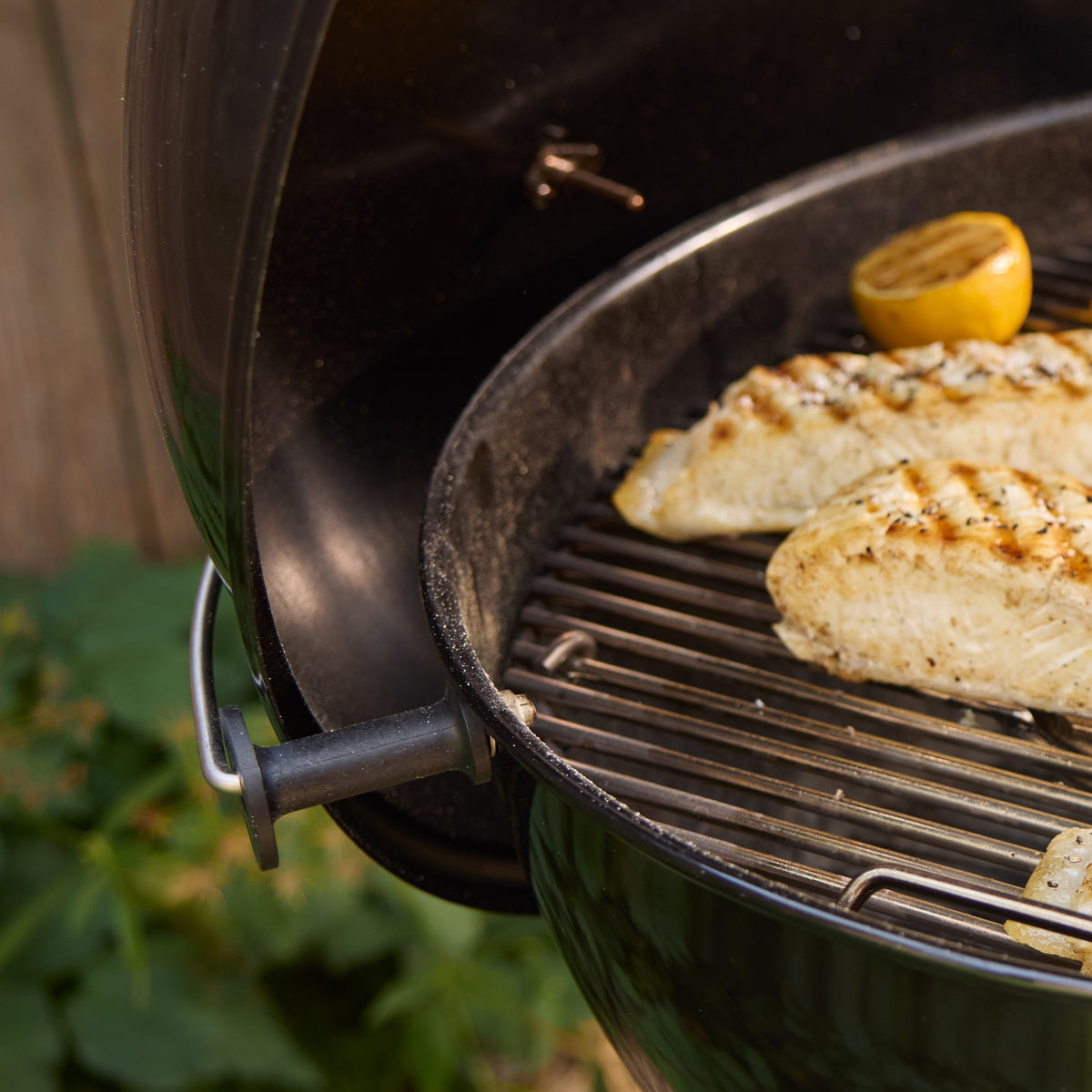 Close-up of a kettle grill’s lid resting in the side lid holder, with fish fillets and a lemon half cooking on the grate.