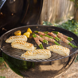 Fish fillets, bacon-wrapped asparagus bundles, and lemon halves cooking on the round grate of an open kettle charcoal grill.