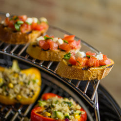 Toasted bread topped with tomatoes, cheese, and basil on the warming rack above stuffed bell peppers on the cooking grate.