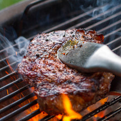 Close-up of tongs gripping a seared steak on the cooking grate, with flames, sparks, and smoke rising around the meat.