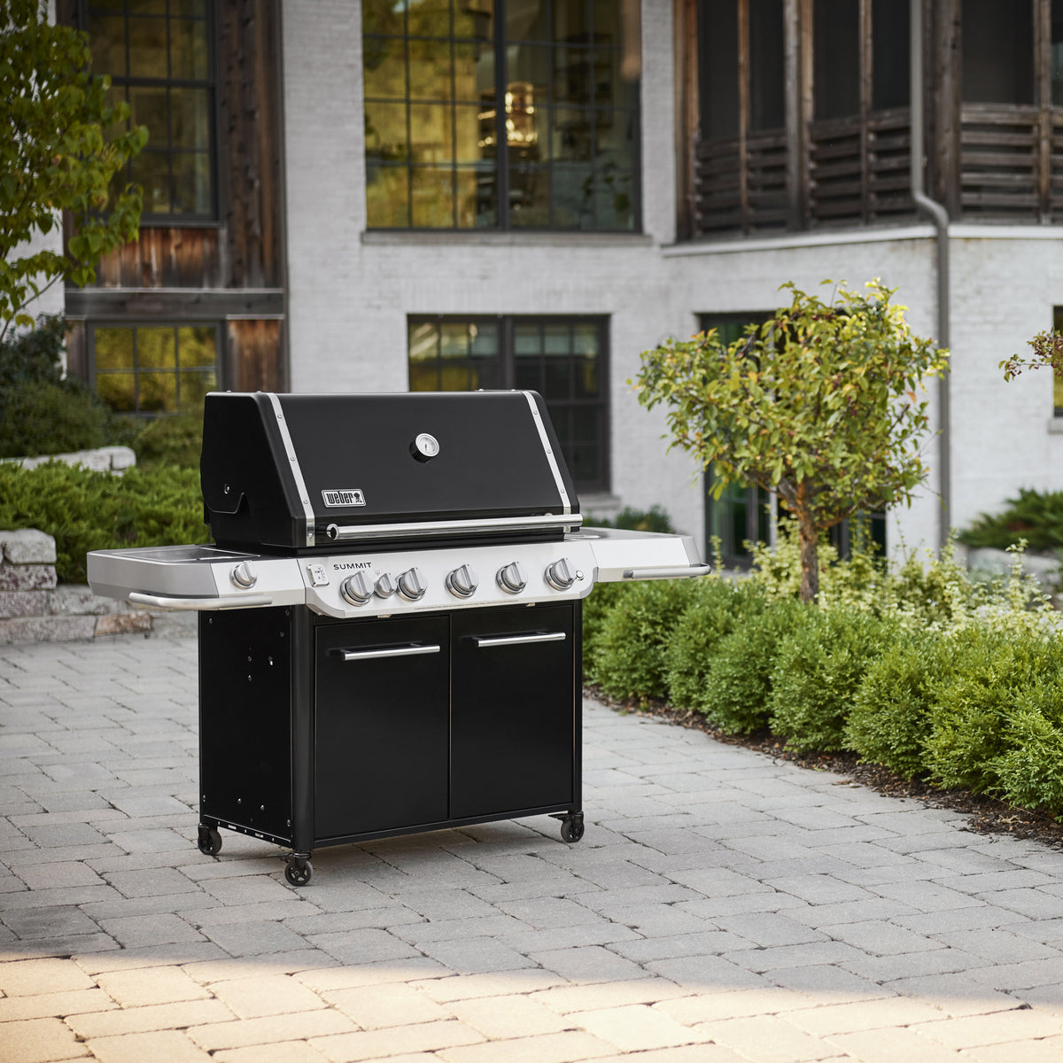 Black Summit FS38 E Gas Grill on a paved patio, with a closed lid, built-in thermometer, stainless-steel side tables, and a two-door cabinet on caster wheels.
