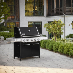 Black Summit FS38 E Gas Grill on a paved patio, with a closed lid, built-in thermometer, stainless-steel side tables, and a two-door cabinet on caster wheels.