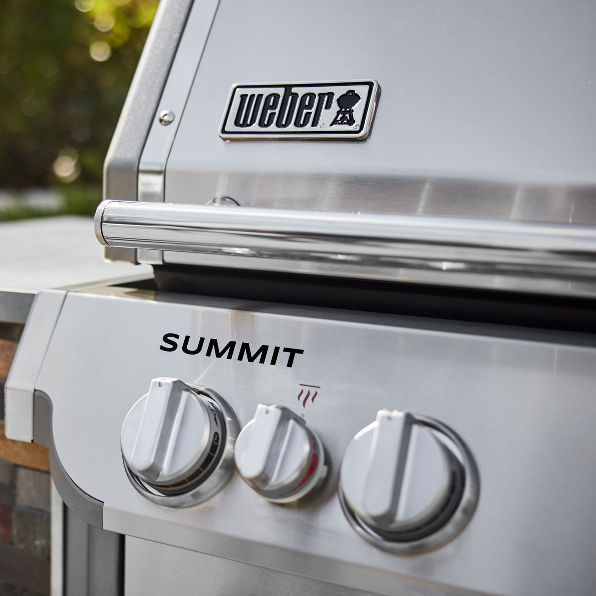 Close-up of a stainless steel gas grill’s lid handle and control panel, with two large burner knobs and a smaller infrared broiler knob marked by a red heat symbol.